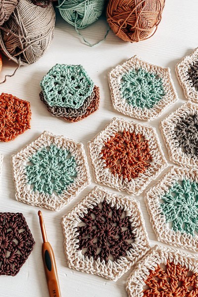 A picture of crochet hexagons in various colours. Brown, aquamarine and orange on a white table with balls of yarn around.