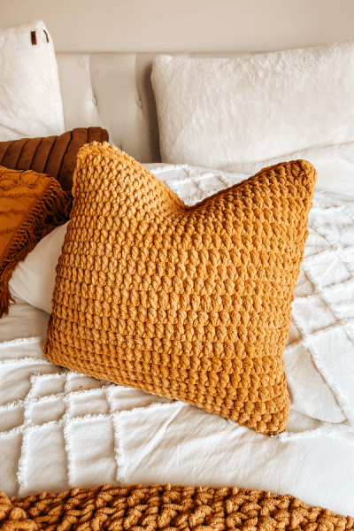 A photo of a crocheted pillow in mustard colour, with texture, on a white bed. The text overlay reads "How to Crochet a Pillow. Free Pattern for Beginners.