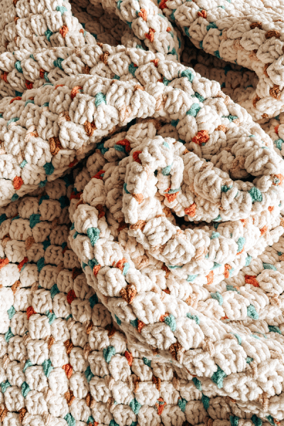 This is an image of a crochet chunky blanket in cream colour with coloured dots. The blanket is twisted in the middle.