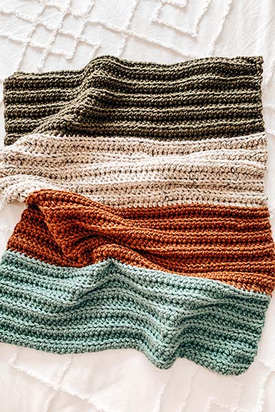 An image of a crochet baby blanket with colour stripes. Green, off white, orange and baby blue on a white bed.